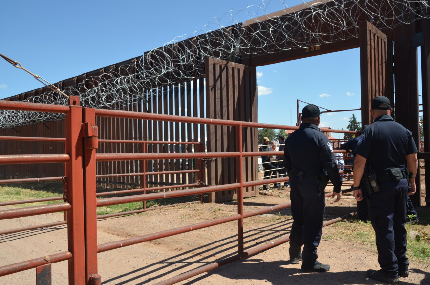 Two people in blue uniforms stand next to orange fence in front of border wall