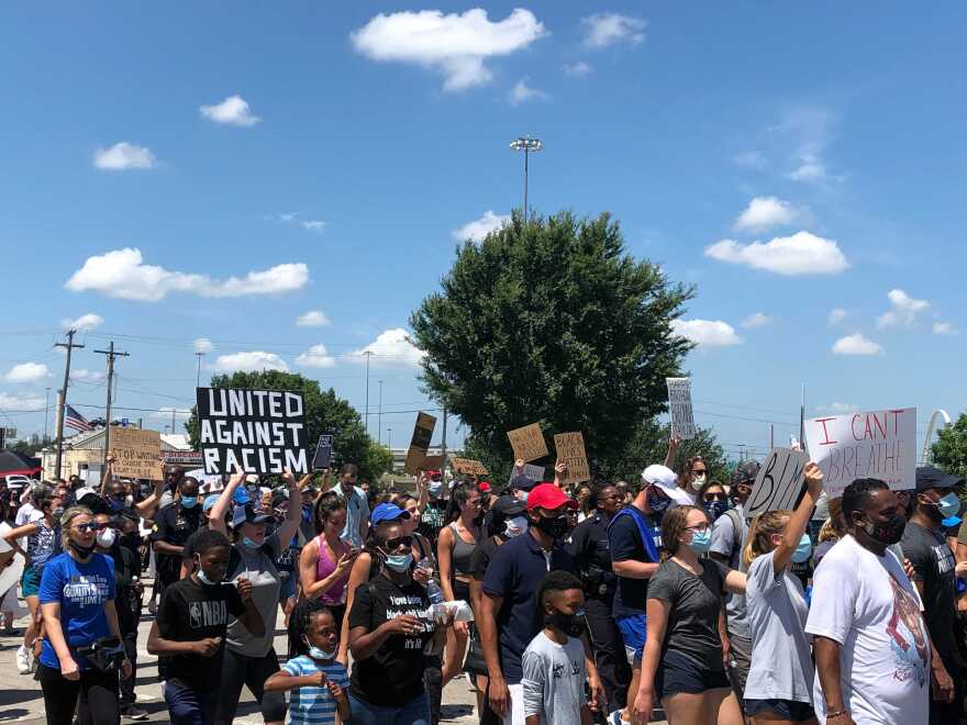 A march of protesters on a summer day, one holding up a sign with a black background and white letters that says "United Against Racism."