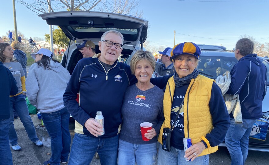 Three smiling adults pose together at a tailgate party. The man and two women wear casual clothes with sports logos. They stand in front of an open SUV, holding drinks, with other fans in the background.