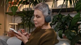 An older woman with short gray hair listens to large headphones with a smartphone in her hand while surrounded by plants.