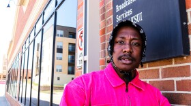 A man in a black bandanna and pink corduroy jacket stands outside of a radio station.