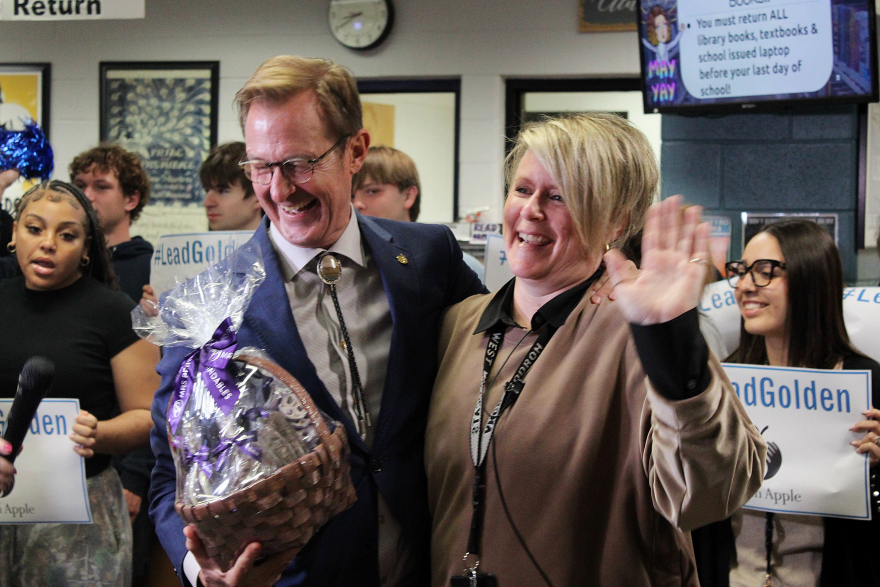 Man in suit hands gift basket to woman who is smiling and waving.