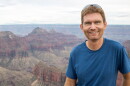 A man with brown hair wears a blue shirt and stands in front of a canyon. He smiles at the camera