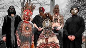 Six people, each person is wearing a different mask. Standing in front of some trees without leaves. The person in the center of the group has a wide legged squat, the rest of the group are either standing or sitting. 