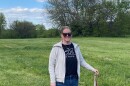 A woman stands in the middle of a grass field and trees behind her.