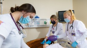 Three nursing students in white scrubs practice CPR on a training dummy.