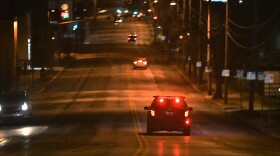 A police cruiser is seen with red lights glowing as it drives down a street at night. There are a few cars on the street, and their tail lights can be seen.