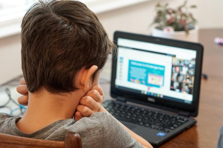 A child with brown hair faces a black laptop with a blurred screen.