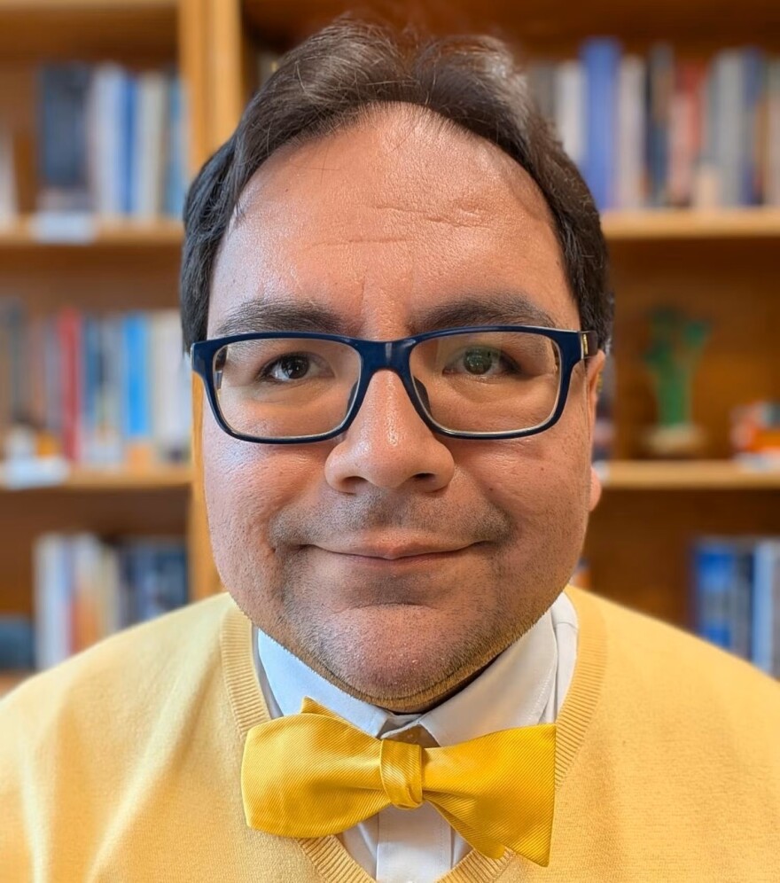 Michaelangelo Landgrave, a man wearing dark-framed glasses, a yellow sweater and a yellow bow tie, smiles for a photo.