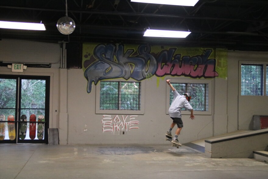 A boy wearing a helmet jumps on a scooter after jumping off a ramp to his right. On the wall in graffiti it reads "Sk8 Church." There's two sets of windows and a set of doors with colorful skateboards leaned up against them.