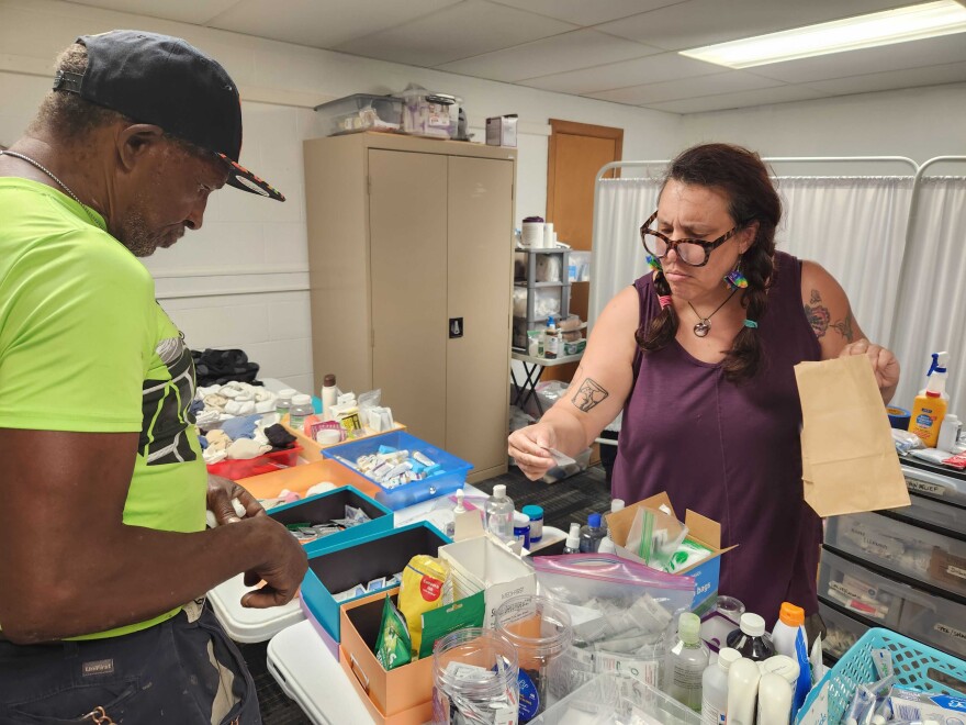 Cat Armbrust, right, is the director of the CoMo Mobile Aid Collective. She is wearing a purple tank top and rainbow earrings. She stands across the table from Bill Tolson who is wearing a ball cap and a bright, neon green shirt. Cat is looking at an anti-itch wipe and holding a brown paper lunch bag.