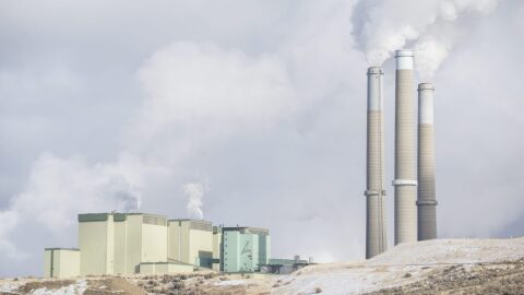 The stacks of a large power plant tower over a hilly snow-covered ground.