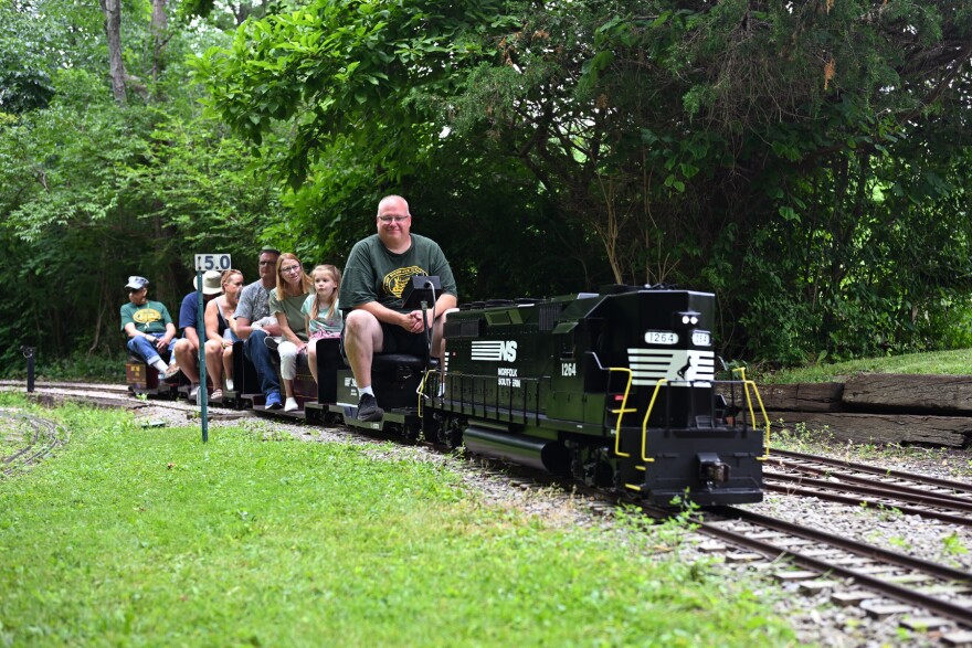 A man drives a miniature train down the tracks with passengers seated behind him