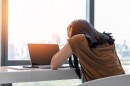 Woman sits at a desk in front of a laptop and holds her head in her hands distressed.
