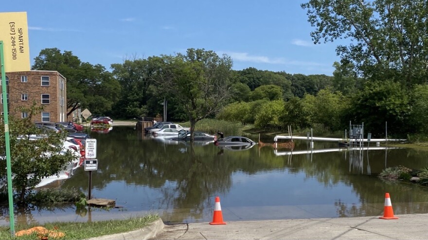 View of flooded parking lot, cones are placed in front of the water. There are at least a dozen cars next to a building partially submerged.