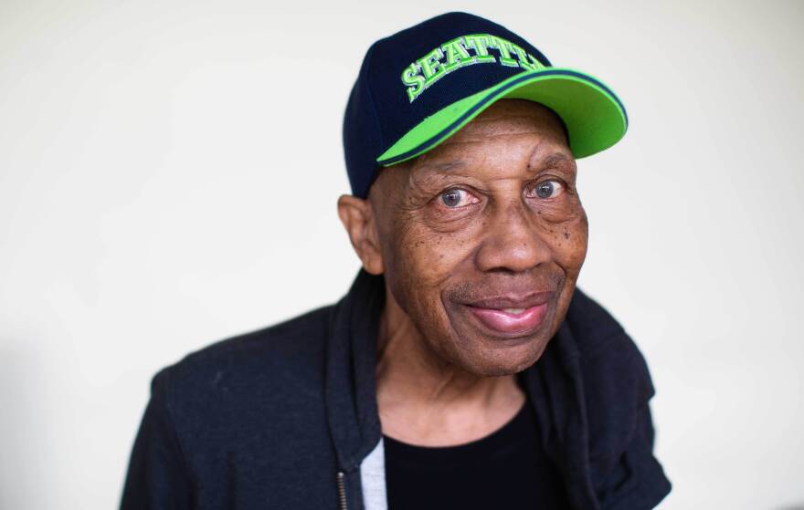 An older Black man half-smiles looking at the camera wearing a Seahawks hat.