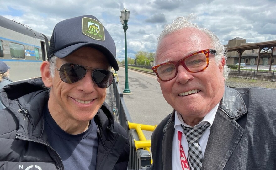 Two smiling men stand outside: a man at left is wearing dark sunglasses, a blue baseball cap, a black puffy jacket, and a blue t-shirt; a man at right has short grey hair and is wearing glasses, a white button-down shirt, a black and white plaid tie; and a black blazer.