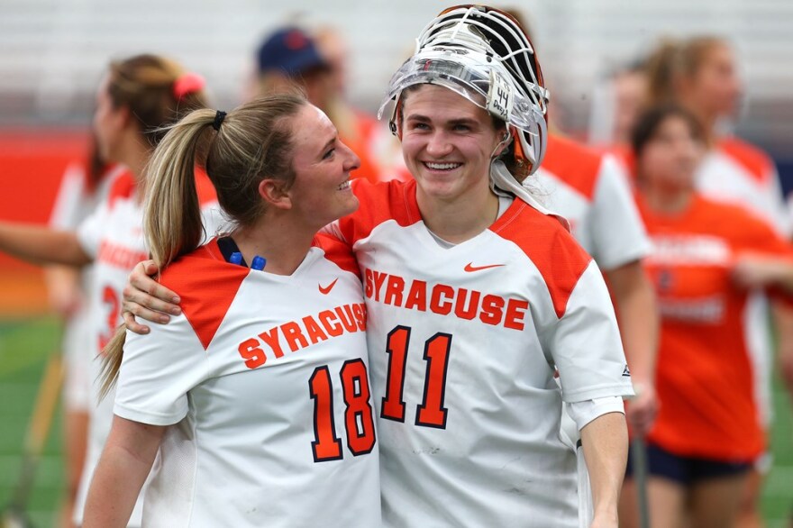 Blonde-haired lacrosse player looks up and smiles at her brunette-haired teammate, both wearing white Syracuse jerseys.