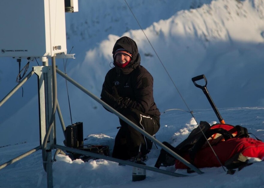 A person bundled up in a snow suit smiles while crouching next to a weather station atop a mountain.