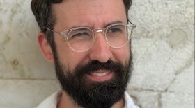A man with a bear and wearing glasses and a white shirt stands against a wall.
