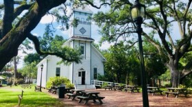 A tall church stands behind large trees and a streetlight. There are steps leading up to the church and benches along the walkway. 