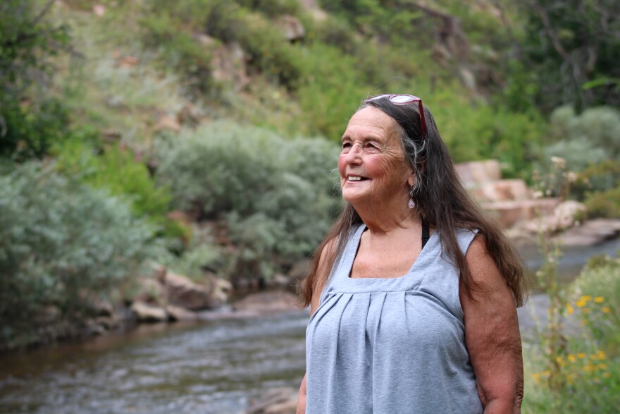 A woman in a blue sleeveless shirt smiles and looks up into the distance. In the background, a river flows through rocks and green bushes.