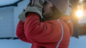 A man in a red coat and large mittens peers through binoculars on a winter morning.