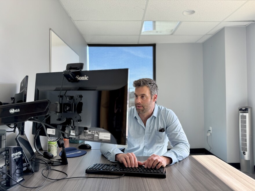 A man in a blue button-down shirt sits at a desk in an office building