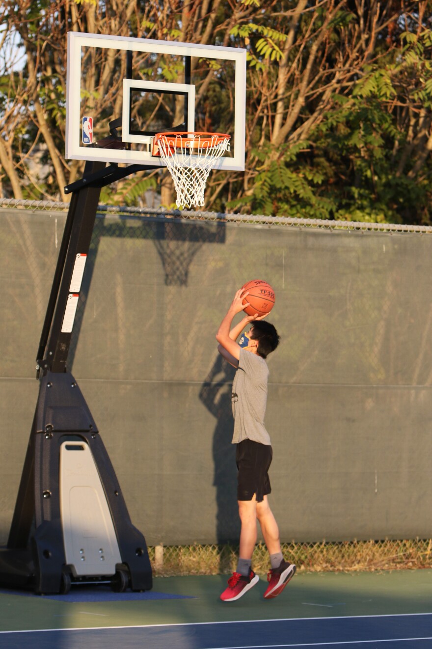 A boy wearing a mask shoots on an outdoor basketball court.