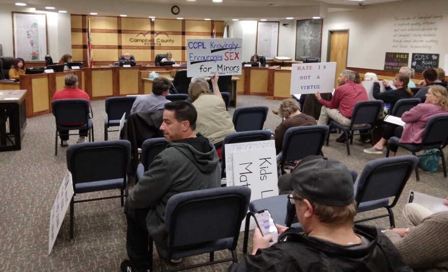 A man amongst a crowd holds a sign that reads "CCPL Knowingly Encourages Sex for Minors" and another that says "Hate is not a virtue" in the Campbell County Commissioner's chamber