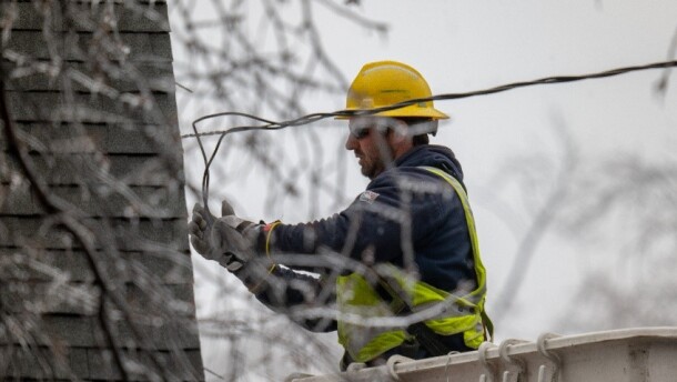 lineman in a cherry picker adjusting wires near a house, there's ice-covered branches out of focus in the foreground
