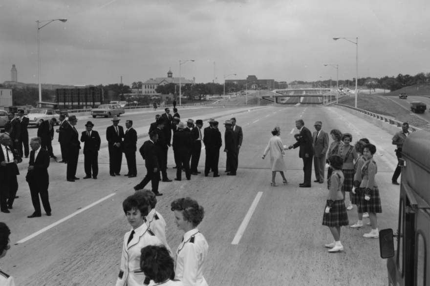 A black and white photo showing crowds standing on I-35 for a dedication on March 29, 1962. Men in black suits mingle. High school girls in school uniforms and wearing white gloves are standing around.