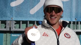 A man in white winter clothes and sunglasses holds up a silver medal.