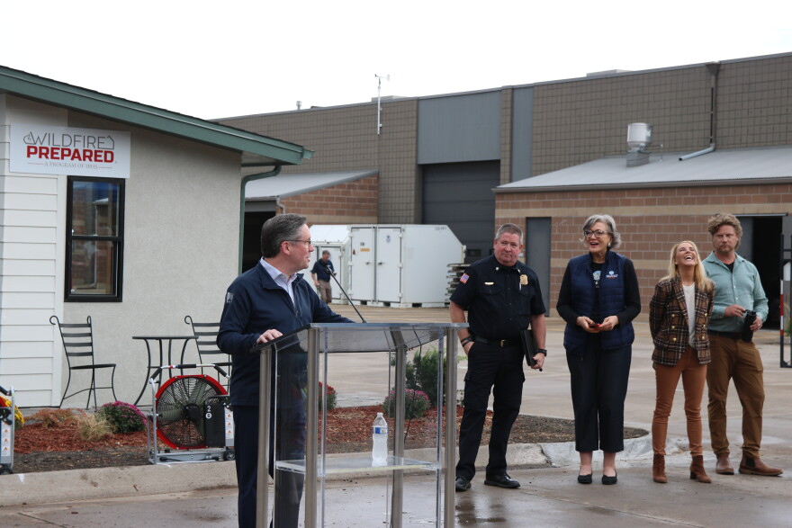 A man with dark hair in a blue polo and dark blue zip-up stands at a clear podium looking back at a line of people that are looking at him. Behind him is a tiny mock home. 