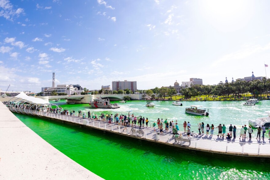 A green river in downtown Tampa with a large group of people standing on a walkway. There is greenery and large buildings in the background. 