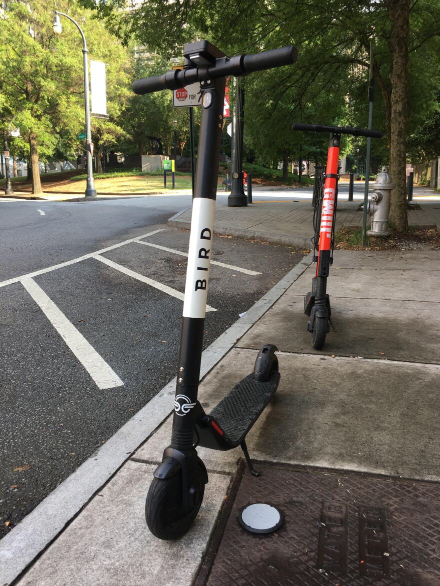 Bird and Jump brand e-scooters lean on their kickstands on the sidewalk. They are faced so that they stand parrallel to the roadway.