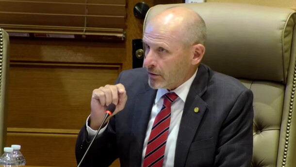 A man in a suit and red striped tie sits at a conference table, speaking into a microphone. He appears to be in a formal meeting room with a wooden door and leather chair visible in the background.