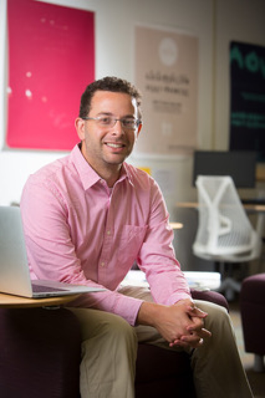 Sam is pictured sitting near a computer, with his elbows on his legs and his hands together. He has brown hair and is wearing glasses, a pink button down, and tan pants.