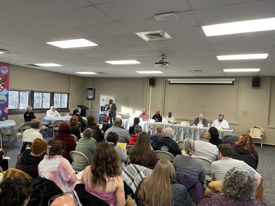 A crowd sits in chairs before people at a table and someone addresses the group, holding a microphone.