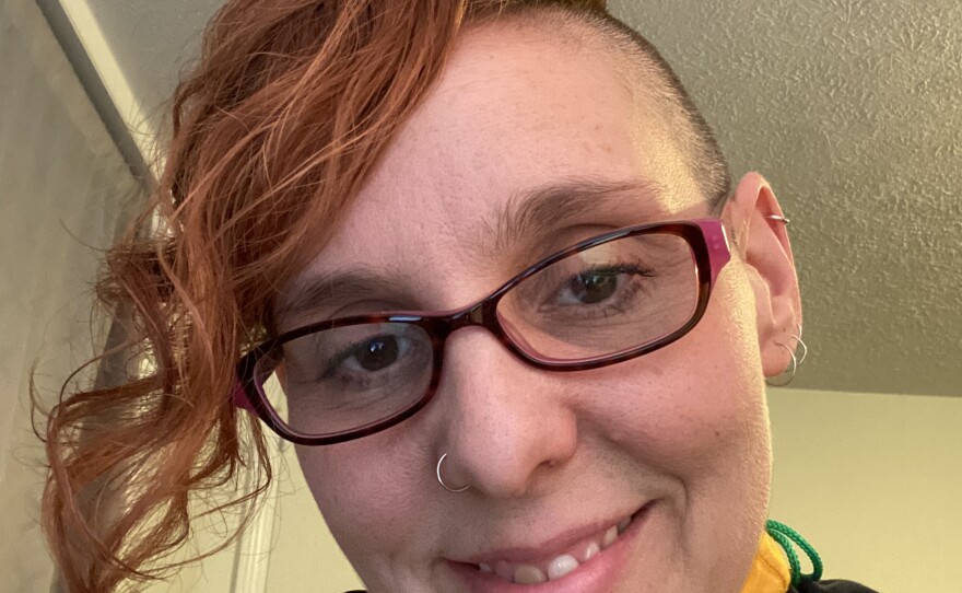 A smiling woman has a partially shaved head with long red hair on one side. She is wearing glasses, a black jacket and a mustard yellow turtleneck.