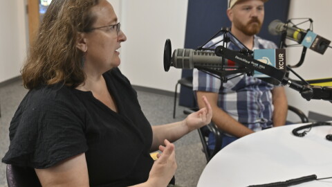 Two people sit in a radio studio talking at microphones. The person at left is gesturing while talking.