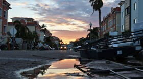 A neighborhood with flooding and debris in the street, and a sunset in the background