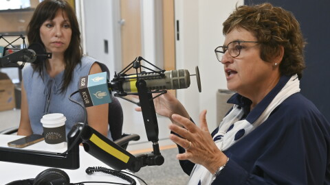 Two people sit in a radio studio talking at microphones. The person at right, Sarah Morris, is gesturing while talking.