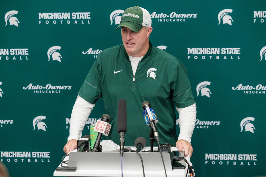 Pat Fitzgerald speaks at a podium with several microphones on it in front of a backdrop with MSU logos. He is wearing a MSU shirt and hat.