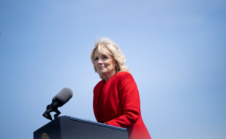 First Lady Jill Biden speaks at a podium, wearing a red jacket, against a bright blue sky.