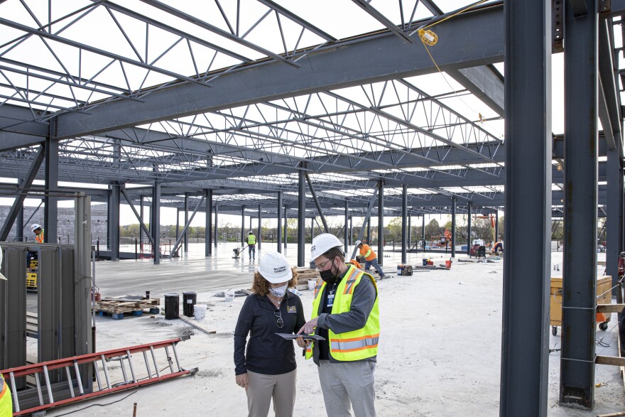 A man and a woman wearing masks and hard hats stand in the middle of a construction site. There is a steel-framed structure without a roof.