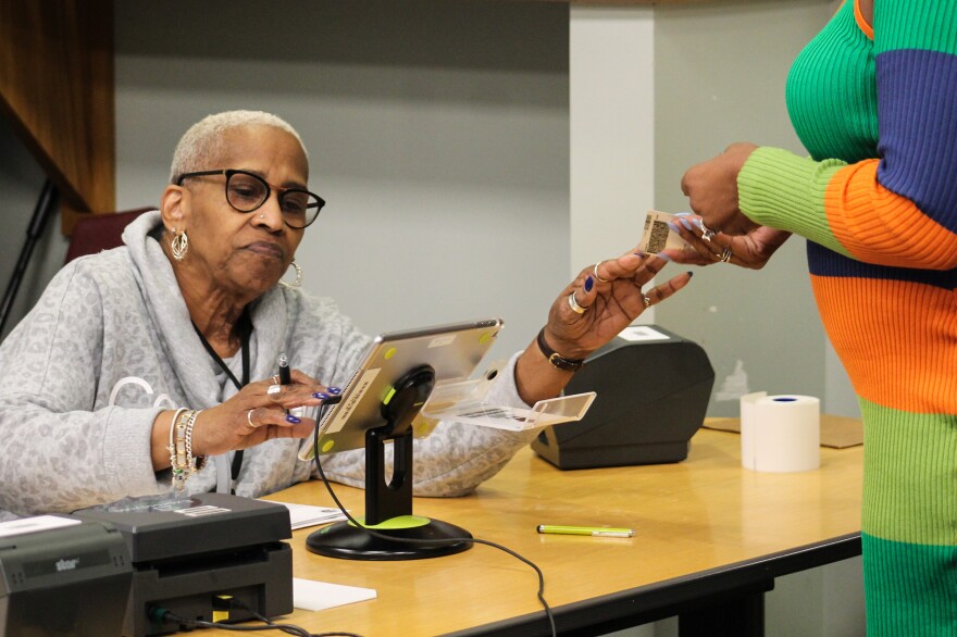 An election worker, an older Black woman with short, white hair and glasses, wearing a gray leopard print top, checks in a voter at a polling place. The election worker is taking the voter's identification card. The voter, shown only from the chest side, side-on, is wearing a brightly-colored dress. 