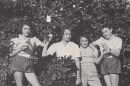 A black and white photograph of four young women posing together in an orchard in front of a leafy citrus tree laden with fruit. They are standing outdoors on small wooden boxes amid tall grass, wearing mid-20th-century casual shirts, shorts, and trousers while holding pieces of fruit.