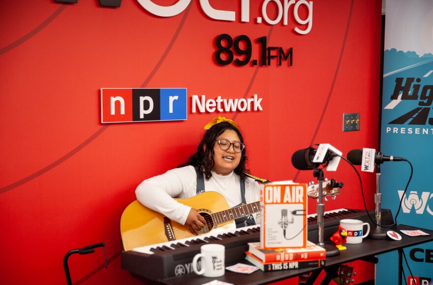 Samara Jane, winner of the 2025 Highway 309 Studio Sessions Contest, performs at WGLT. She sits in front of a red WGLT and NPR Network backdrop, playing an acoustic guitar and singing into a studio microphone, with a keyboard and radio gear on the table in front of her.