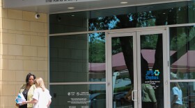Two people stand outside of JFS Dallas' new Addison campus. A grey, metal awning is above a set of double glass doors with the JFS Dallas Logo.
